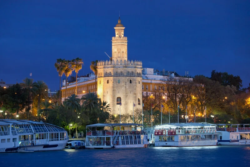 Torre del Oro iluminada à noite com barcos no Rio Guadalquivir em Sevilha, um dos passeios mais encantadores para quem procura o que fazer em Sevilha