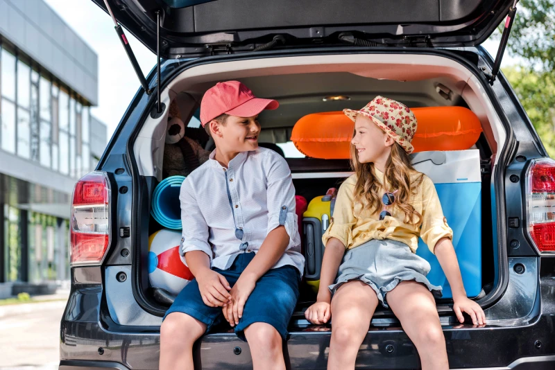 Duas crianças sentadas no porta-malas de um carro, prontas para viagem com crianças no Brasil nas férias escolares.