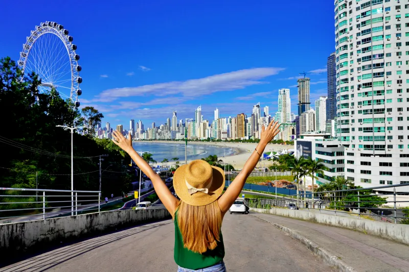 Turista comemorando as férias em Balneário Camboriú com vista para a roda-gigante, praia e arranha-céus ao fundo