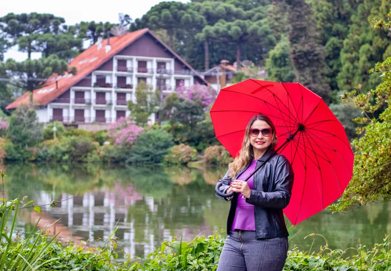 Lago Negro em Gramado com Carol do Vamos Por Aí segurando guarda-chuva vermelho diante de casas em estilo enxaimel.